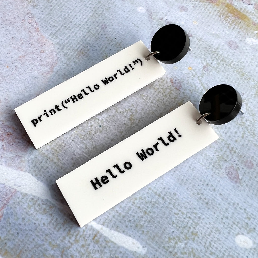White dangle asymmetrical acrylic earrings with black text showing code to display Hello World. Hanging from black earring toppers .