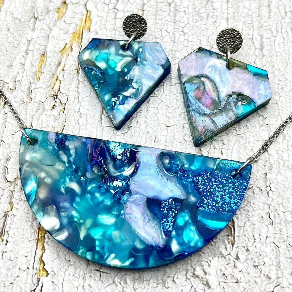 A matching necklace and earrings set made from blue toned acrylic scrap. The necklace is a semi-circle and the earrings are diamond shaped.