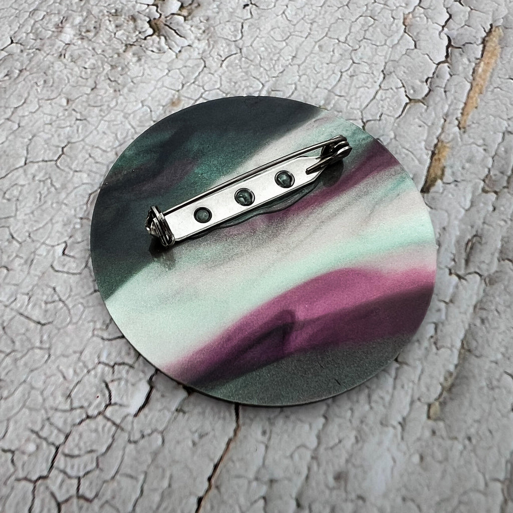 The back of a round acrylic brooch, showing the stainless steel brooch pin and rolling clasp.
