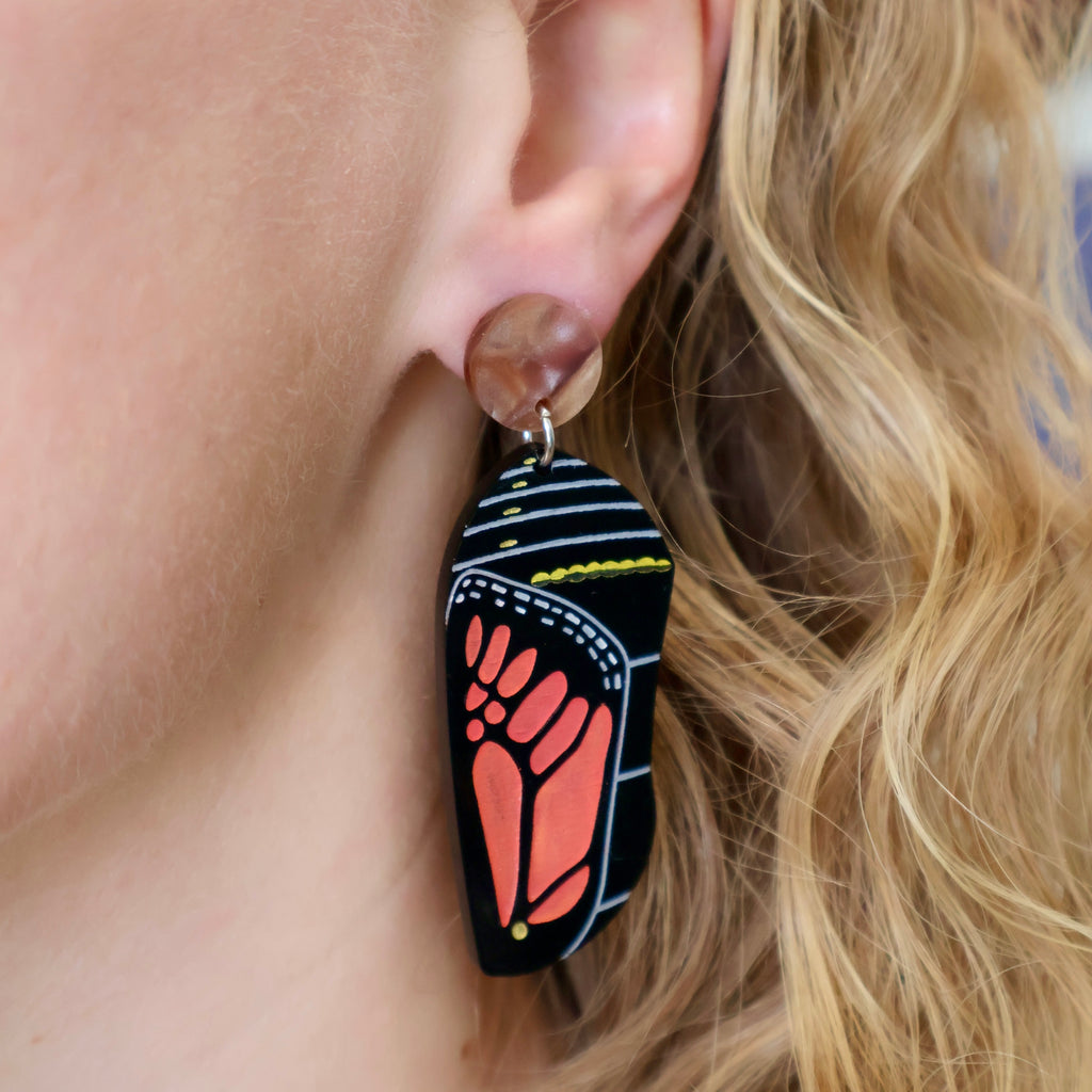 a laser cut acrylic monarch butterfly chrysalis earring being modelled. Laser cut and handpainted from acrylic. Design shows the late stage of the chrysalis in black and red tones. 