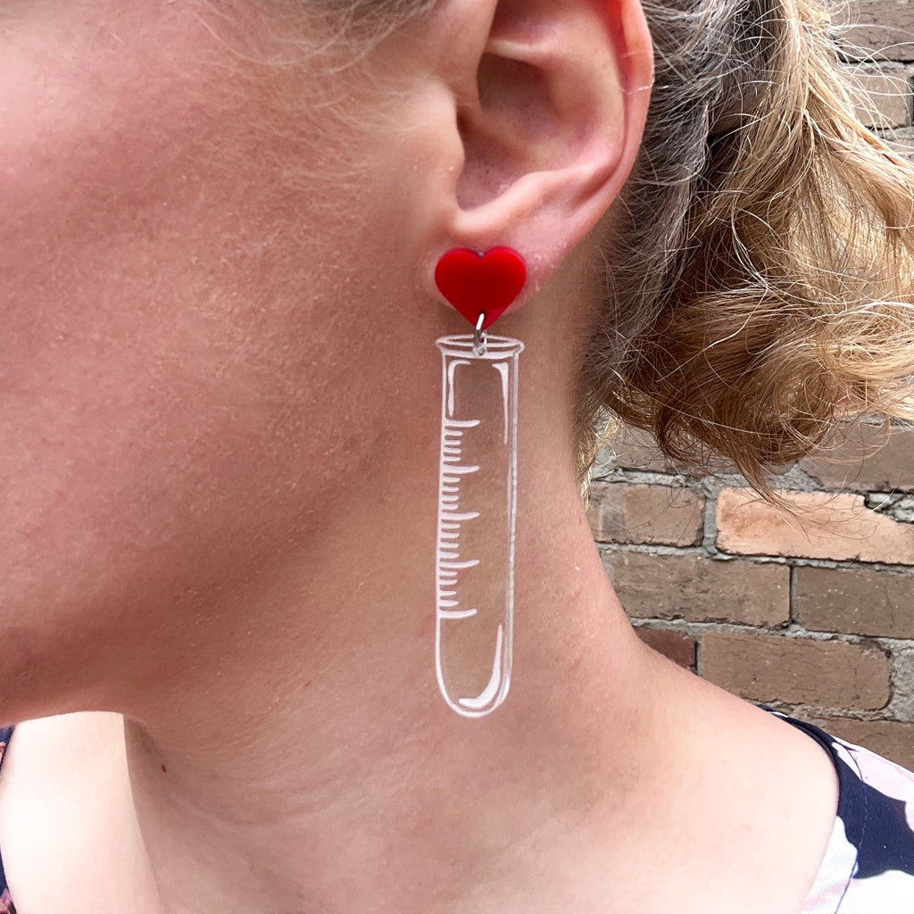 Test tube earrings, laser cut and engraved in clear acrylic, and hanging from red heart earring toppers. Earrings are being modelled. 