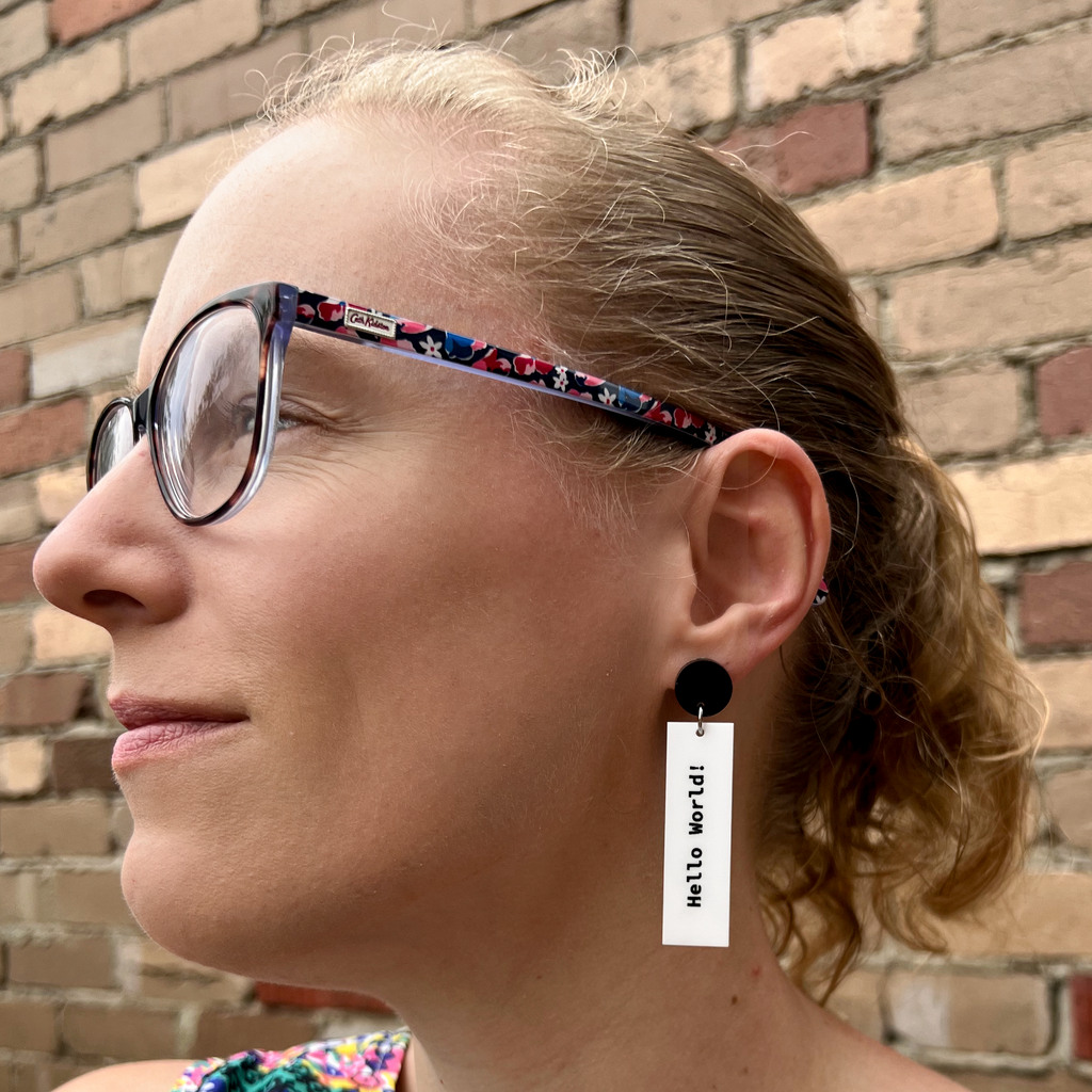 Rectangular white dangle earrings with print("Hello World!")  in black text on one side and 'Hello World!' on the other side. They hang from black acrylic toppers. Earrings are being modelled. 