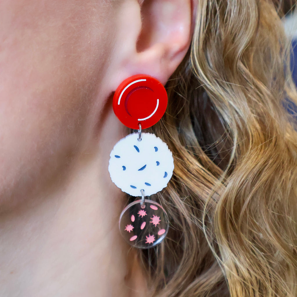 Dangle earrings comprising of a red blood cell topper, with a white blood cell and platelets dangling underneath. Laser cut and handpainted from acrylic. Earrings are being modelled. 