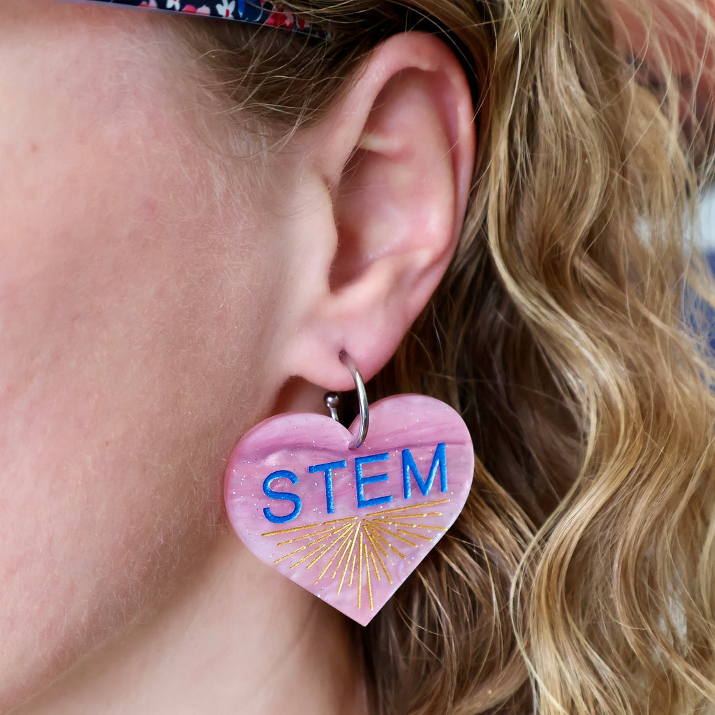 Swirly pink acrylic heart earrings with blue STEM text and a gold starburst pattern, hanging from stainless steel toppers. Earrings being modelled.