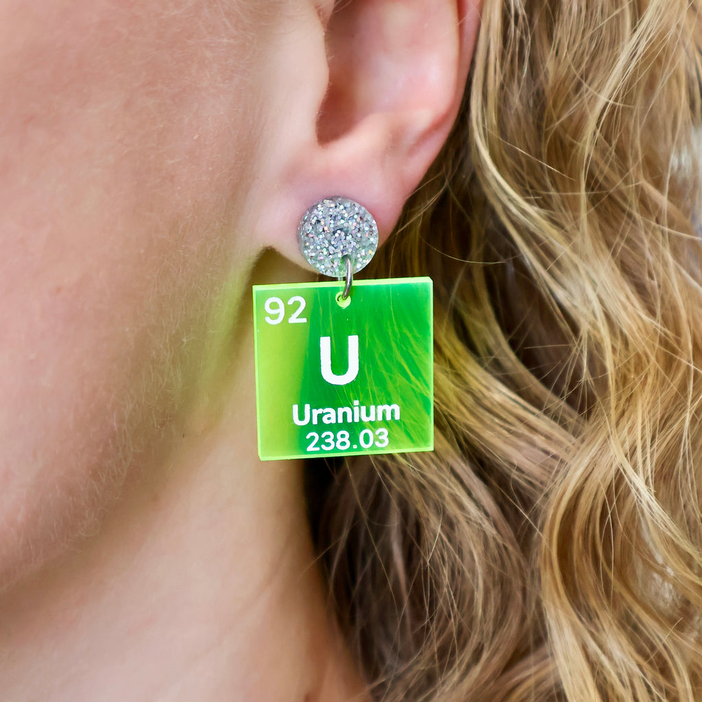 Uranium periodic table earrings, with white text on bright yellow transparent acrylic squares, hanging from silver glitter toppers. Earrings being modelled.