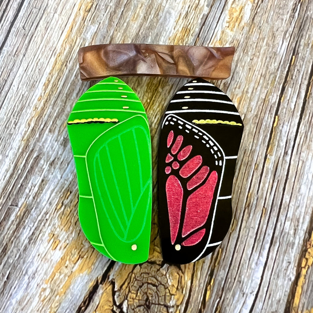 A brooch showing two stages in a monarch butterfly chrysalis development, with one side being a green chrysalis with the other being a late stage black and red chrysalis, both hanging from a brown acrylic ‘branch’.