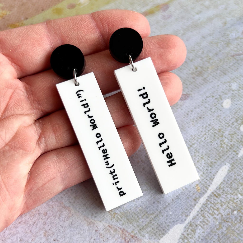 Closeup view of rectangular white dangle earrings with print("Hello World!")  in black text on one side and 'Hello World!' on the other side. They hang from black acrylic toppers. 