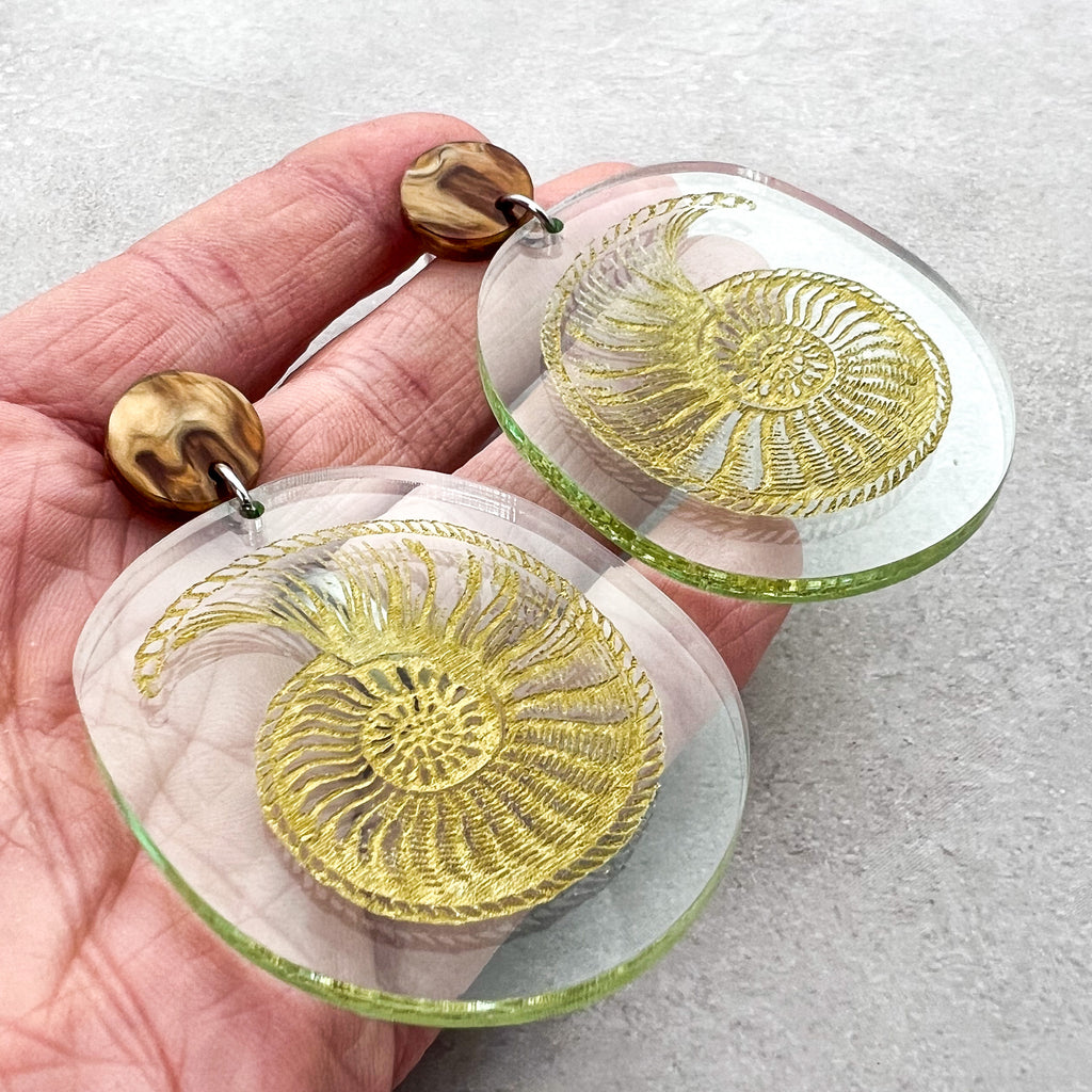 Nautilus fossil earrings, with a gold nautilus design handpainted on a green seaglass toned acrylic. They earrings hang from brown acrylic toppers. 
