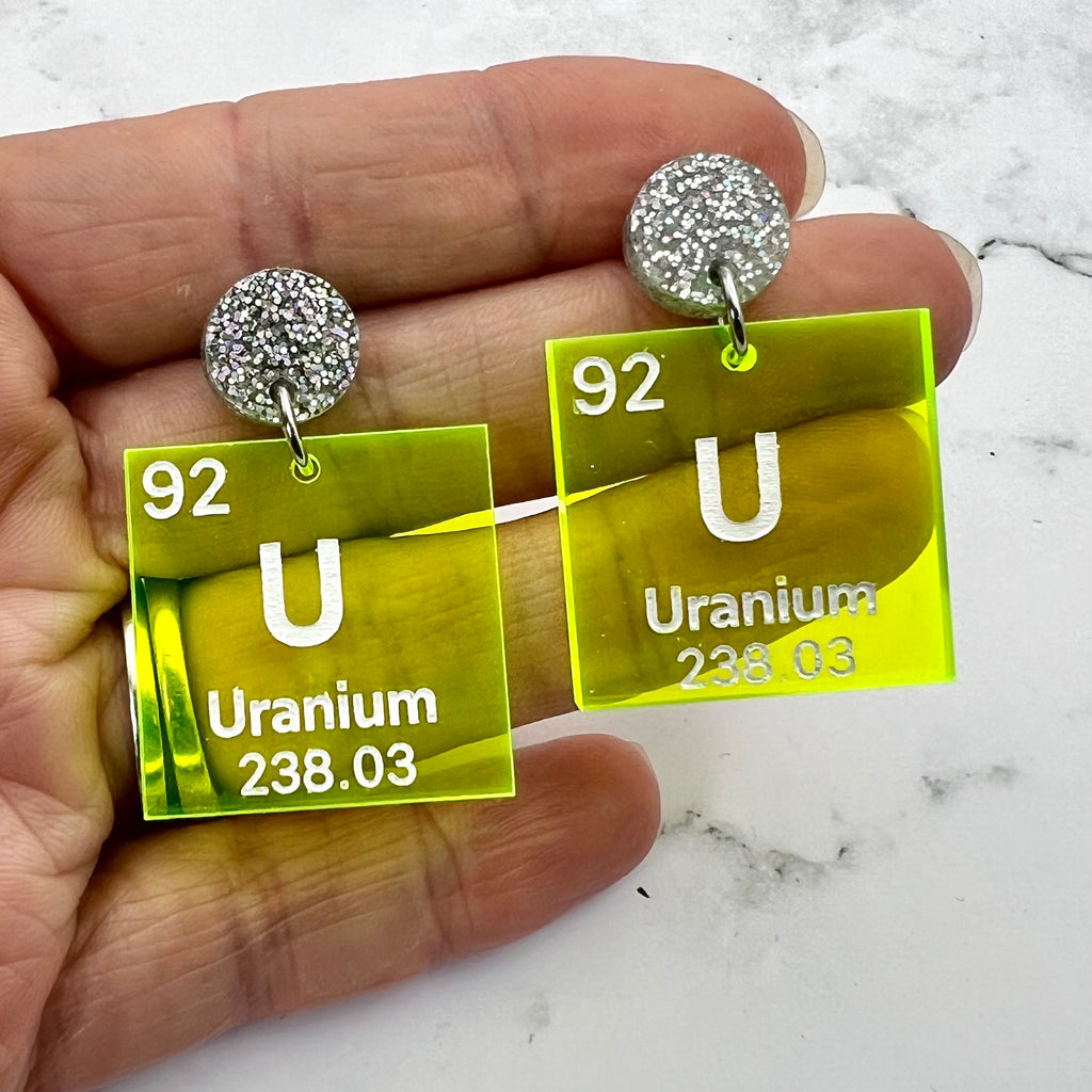 Uranium periodic table earrings, with white text on bright yellow transparent acrylic squares, hanging from silver glitter toppers. Closeup view.