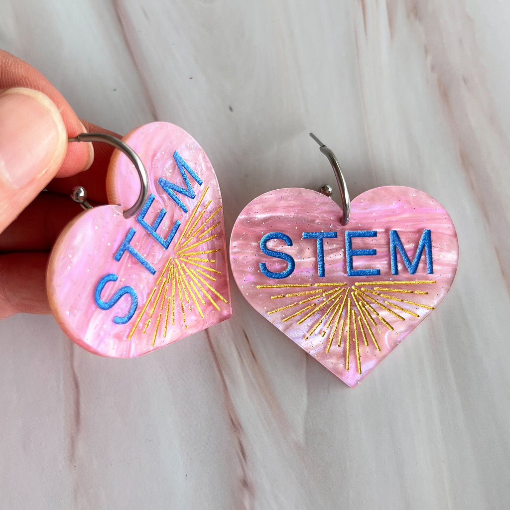 Swirly pink acrylic heart earrings with blue STEM text and a gold starburst pattern, hanging from stainless steel toppers. Angled view.