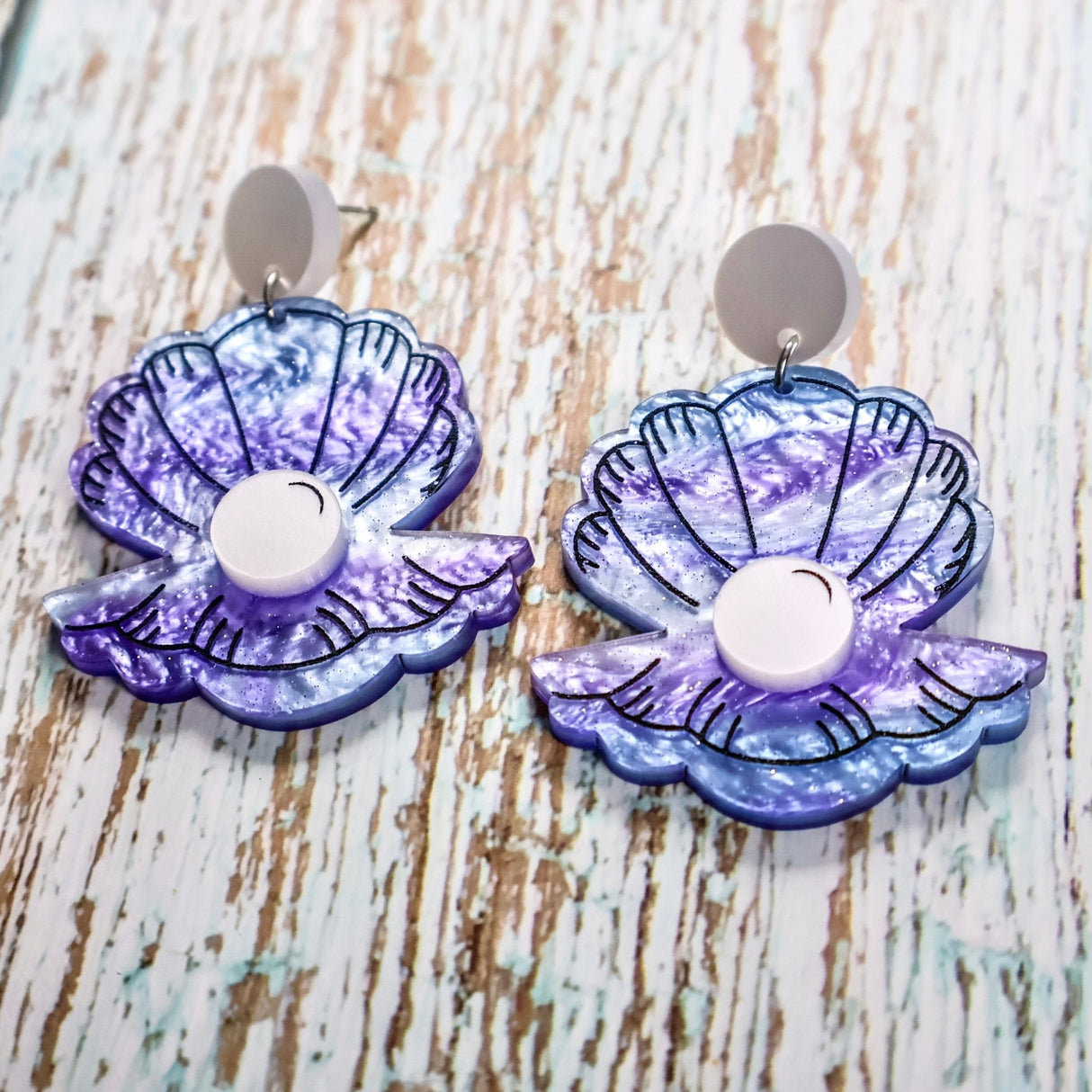 Pair of purple and blue acrylic clam and pearl earrings on a wooden background. Acrylic is swirly with glitter accents. The 'Pearl' is laser cut in a pearlescent acrylic.
