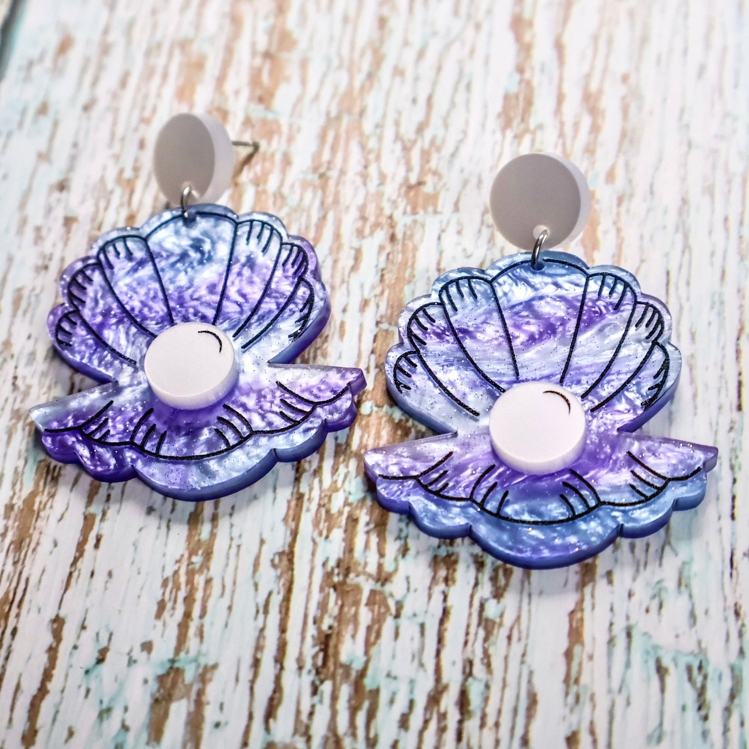 Pair of purple and blue acrylic clam and pearl earrings on a wooden background. Acrylic is swirly with glitter accents. The 'Pearl' is laser cut in a pearlescent acrylic.