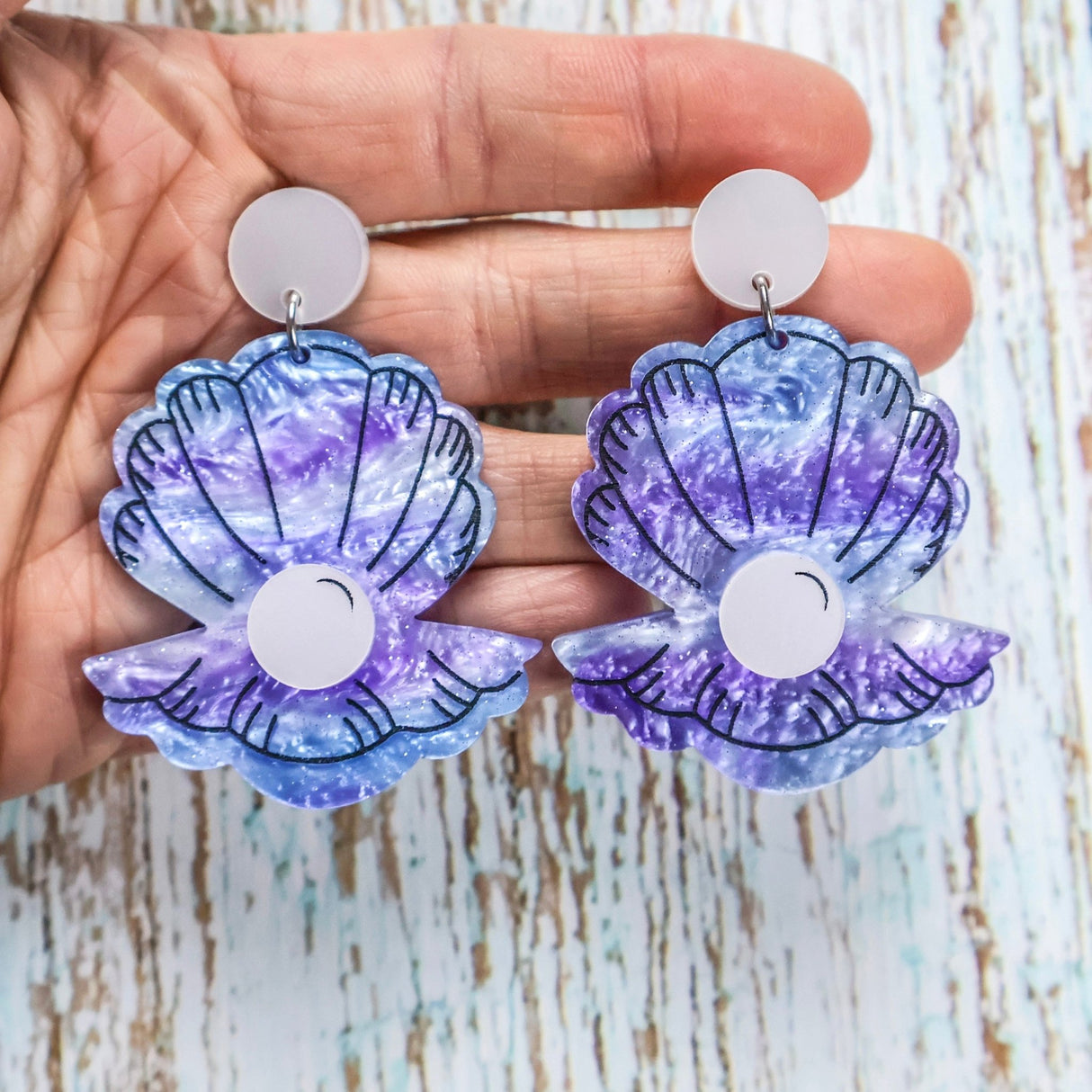 Pair of purple and blue acrylic shell-shaped earrings held in a hand against a wooden background.