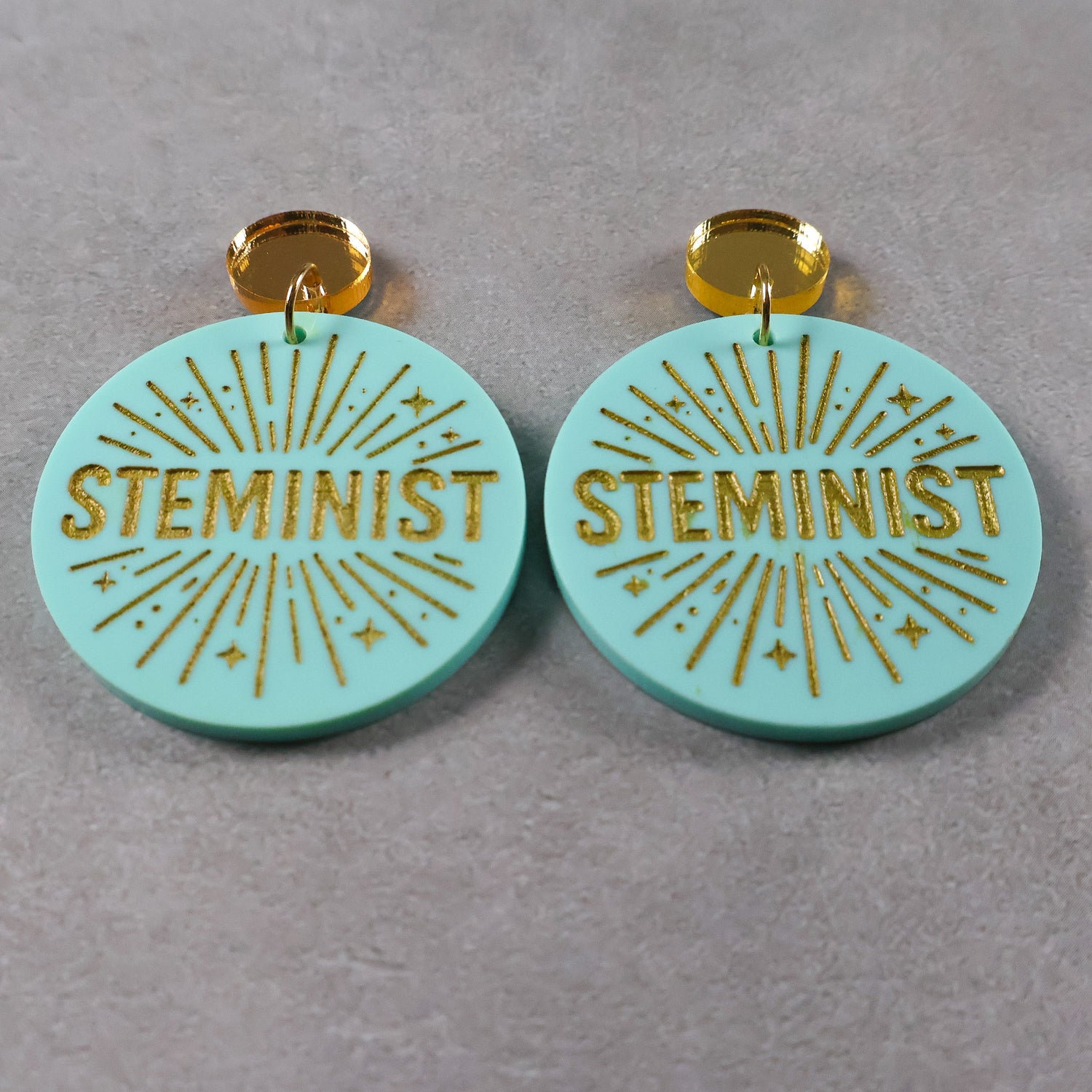 Turquoise or teal dangle earrings with 'STEMINIST' gold text and gold mirror acrylic toppers