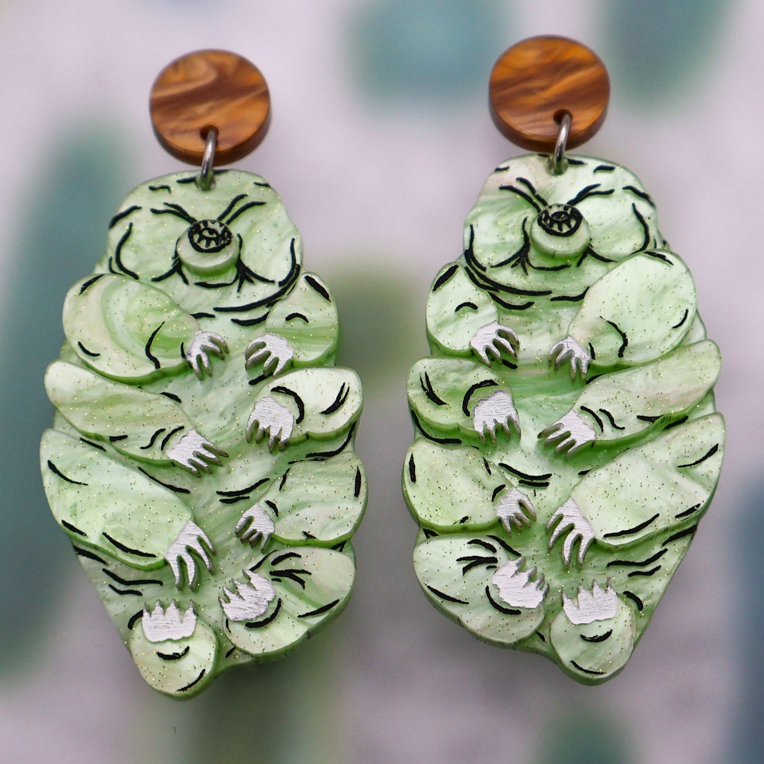 Tardigrade earrings made from swirly apple green acrylic, hanging from swirly brown round earring toppers. Handpainted details and multilayered design.