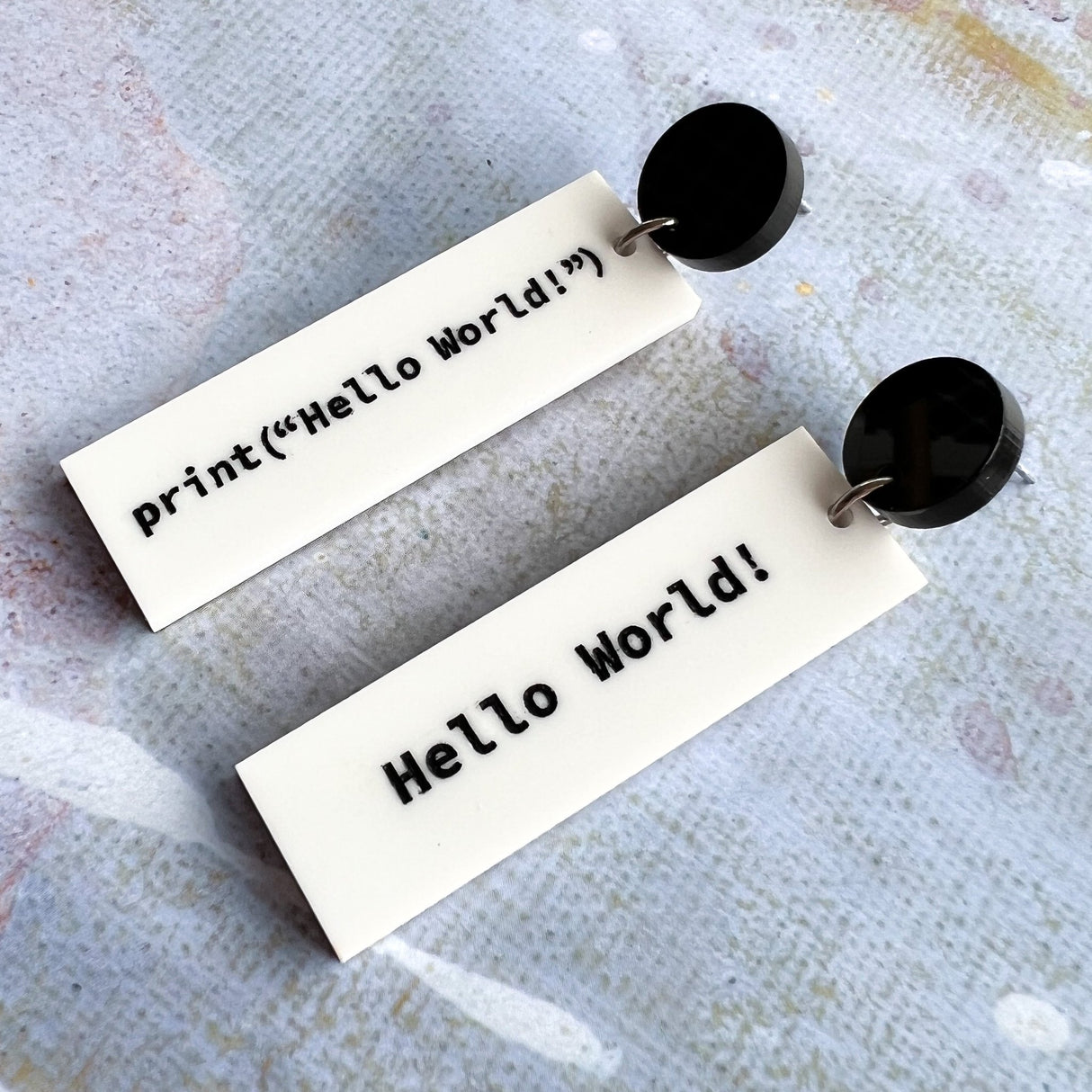 Rectangular white dangle earrings with print("Hello World!")  in black text on one side and 'Hello World!' on the other side. They hang from black acrylic toppers.