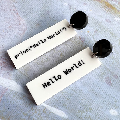 Rectangular white dangle earrings with print("Hello World!")  in black text on one side and 'Hello World!' on the other side. They hang from black acrylic toppers.