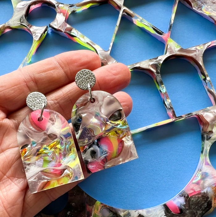 Colourful arch shaped earrings held in a hand with a blue background and the leftovers of a recycled acrylic sheet.