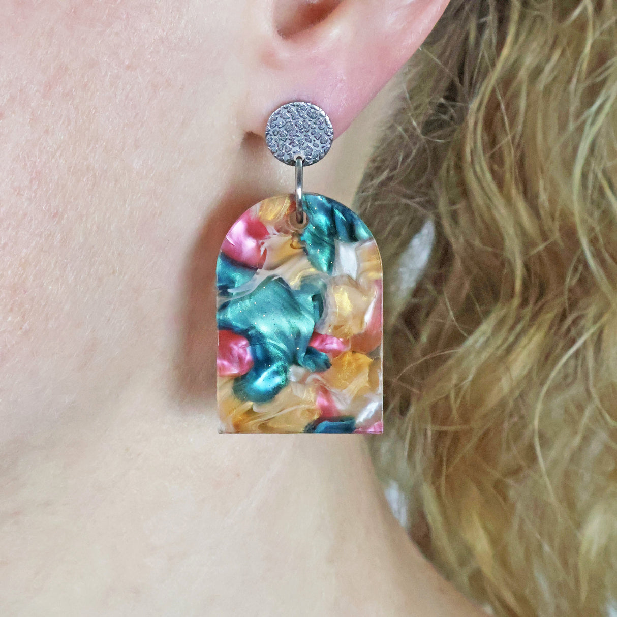 Arch shaped recycled acrylic earrings  in pink, green and caramel tones with stainless steel toppers being modelled on the ear of a person with blonde hair.