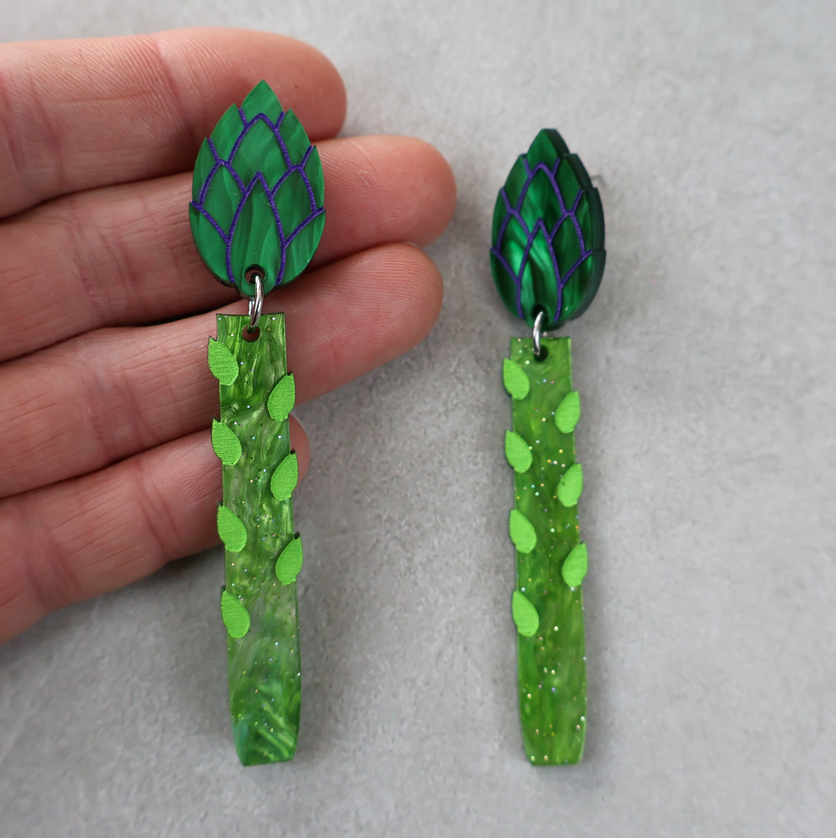 Green asparagus acrylic earrings with leaf-shaped toppers earrings held by a hand on a grey background