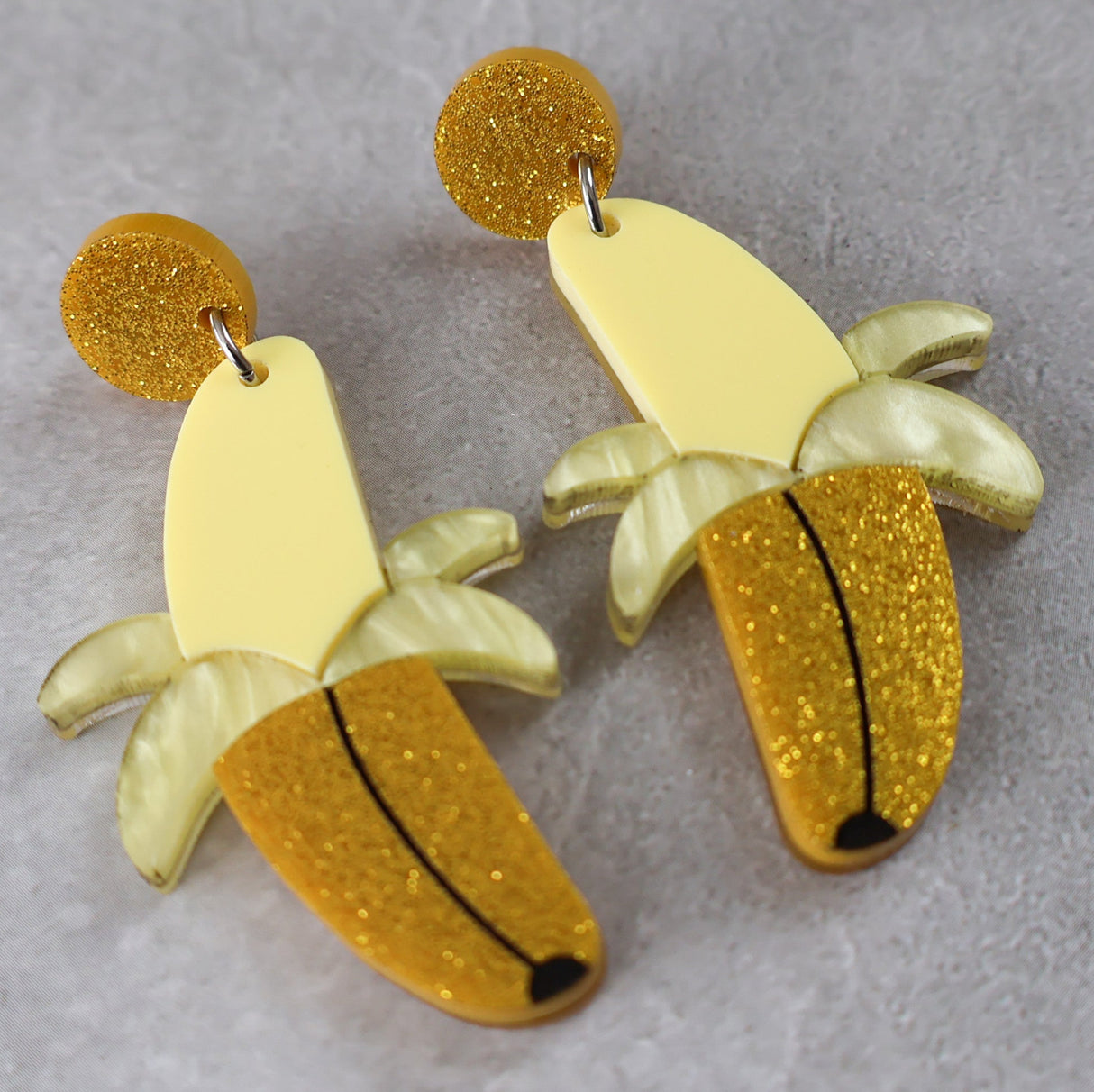 Banana-shaped earrings half peeled with yellow and gold glittery finishes on a grey background