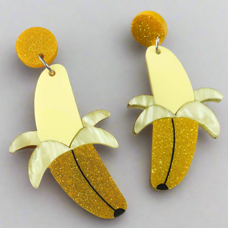 Banana-shaped earrings half peeled with yellow and gold glittery finishes held by a hand on a light grey background.