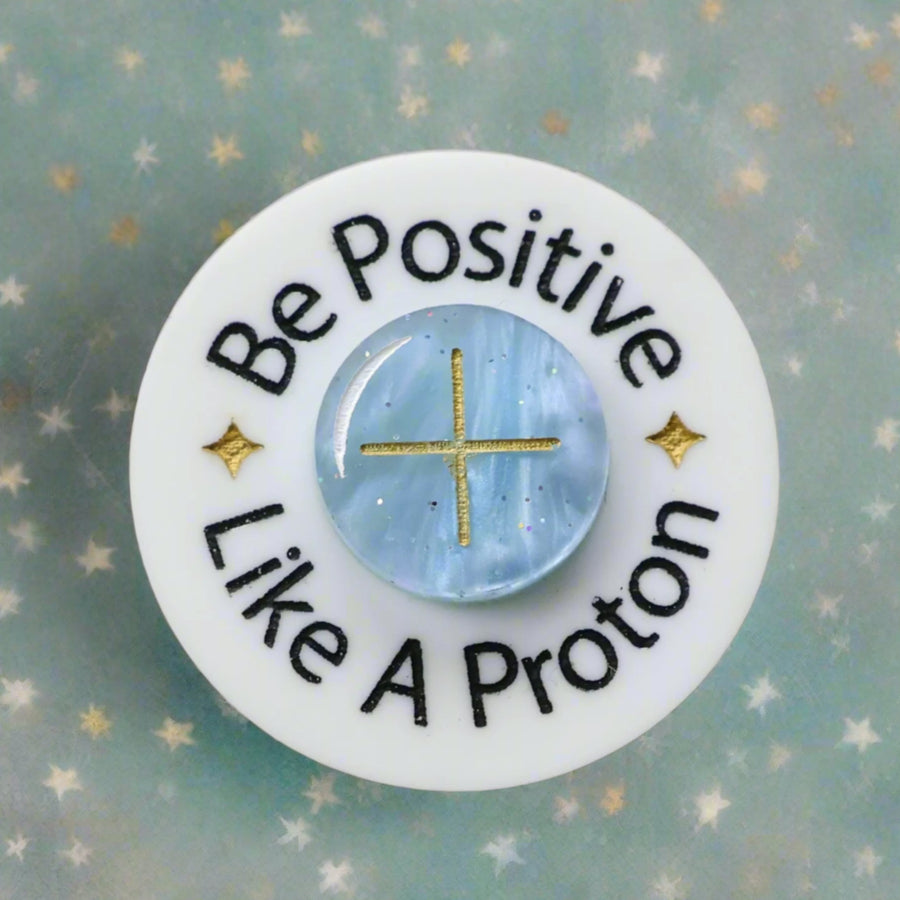 Circular brooch with 'Be Positive Like A Proton' text and a blue 'proton' in the centre