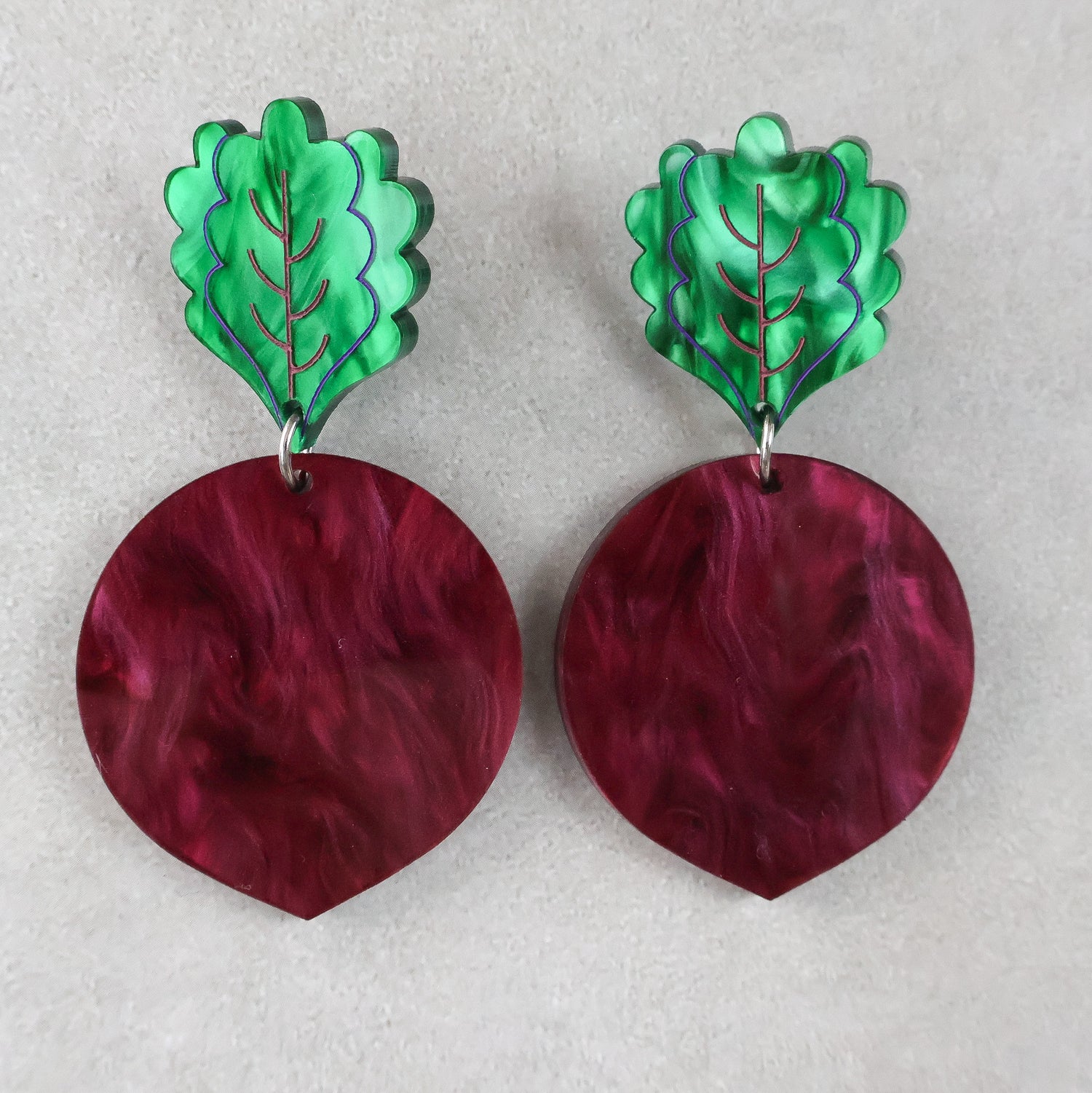 Pair of acrylic beetroot earrings with green leaf-shaped tops and marbled maroon circular bodies on a light grey background