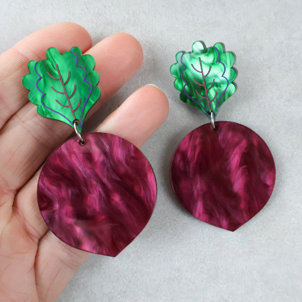 Pair of acrylic beetroot earrings with green leaf-shaped tops and marbled maroon circular bodies held by a hand on a light grey background