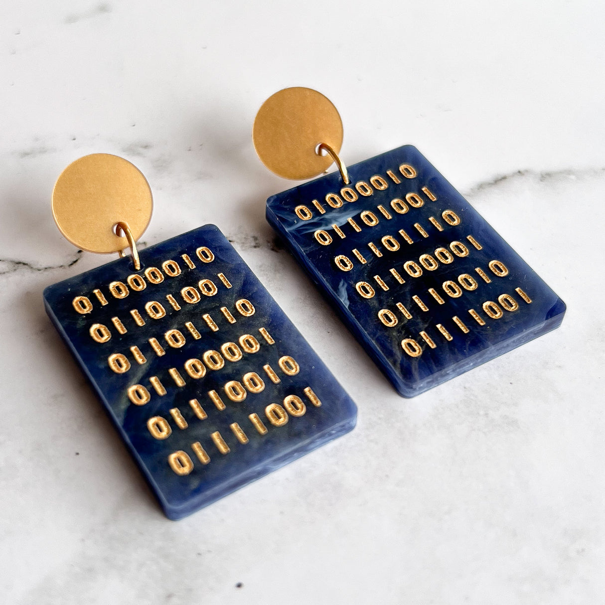 A pair of blue rectangular acrylic earrings with gold binary text engraved within, hanging from gold toned acrylic toppers.