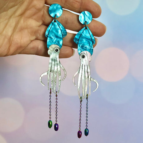 Blue toned squid-shaped earrings held in a hand with chain dangling off the bottoms.