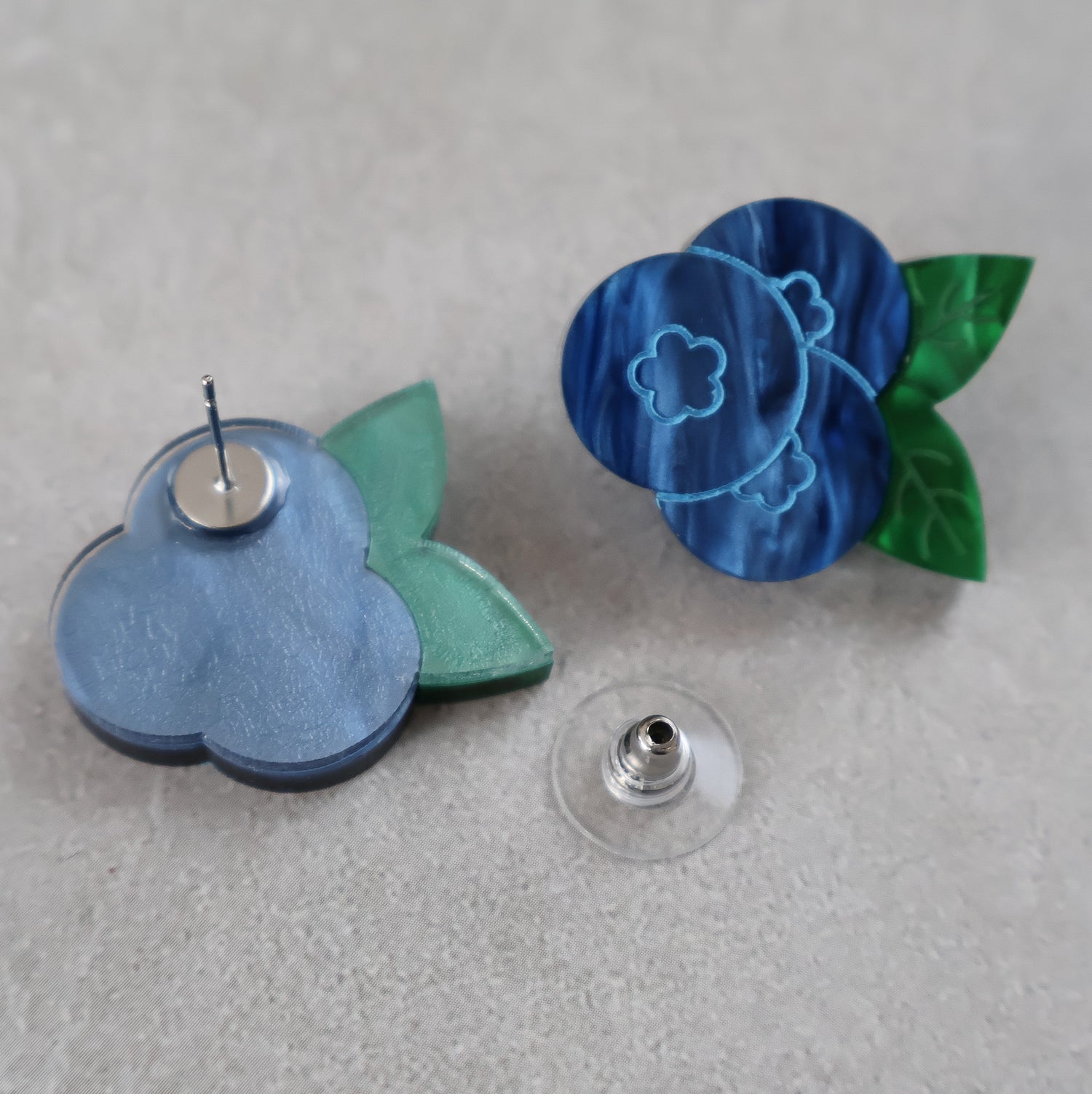 Blue and green blueberry acrylic earrings on a gray surface. One earring is turned to show the stainless steel earring post.