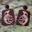 Pair of brown earrings with dinosaur skeleton design on a wooden surface with a fern nearby