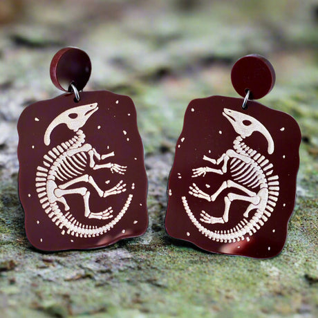 Pair of brown earrings with dinosaur skeleton design on a wooden surface with a fern nearby
