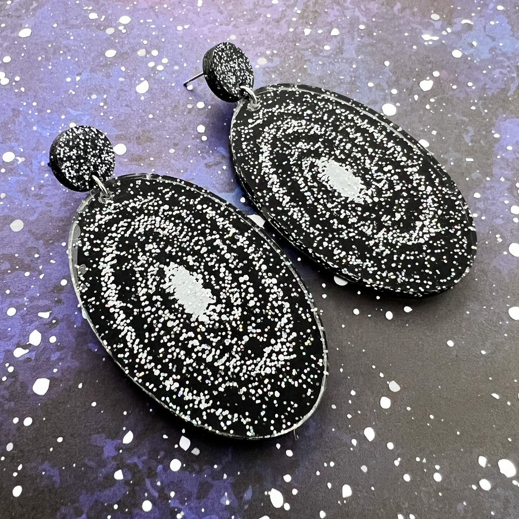Statement sized milky way galaxy acylic earrings. The earrings are oval in shape and contain silver toned stars handpainted against a black background. The earrings hang from black glitter acrylic toppers. 