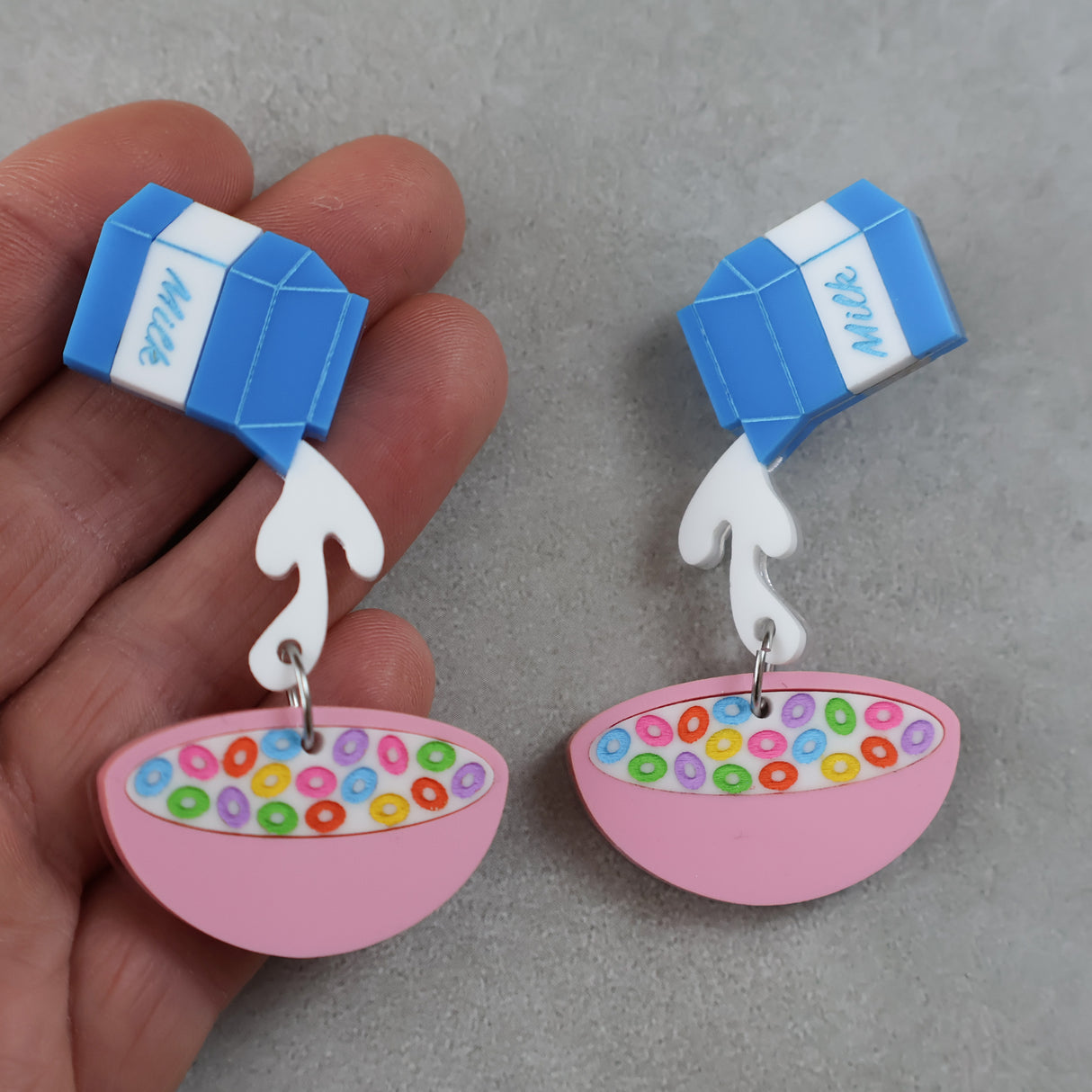 Milk carton and cereal bowl acrylic earrings held by a hand on a grey background