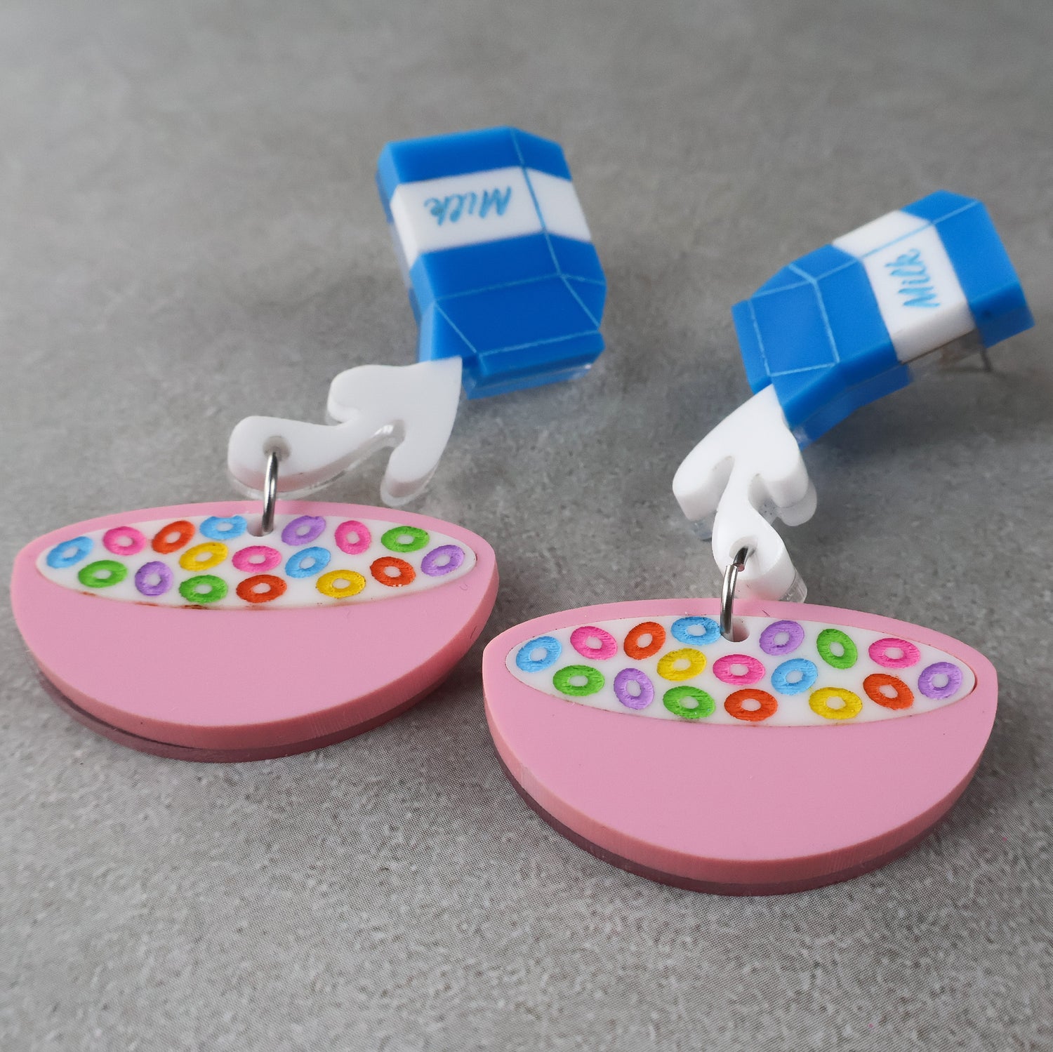 Closeup view of a pair of acrylic earrings shaped like a milk carton pouring into a bowl of cereal on a gray background