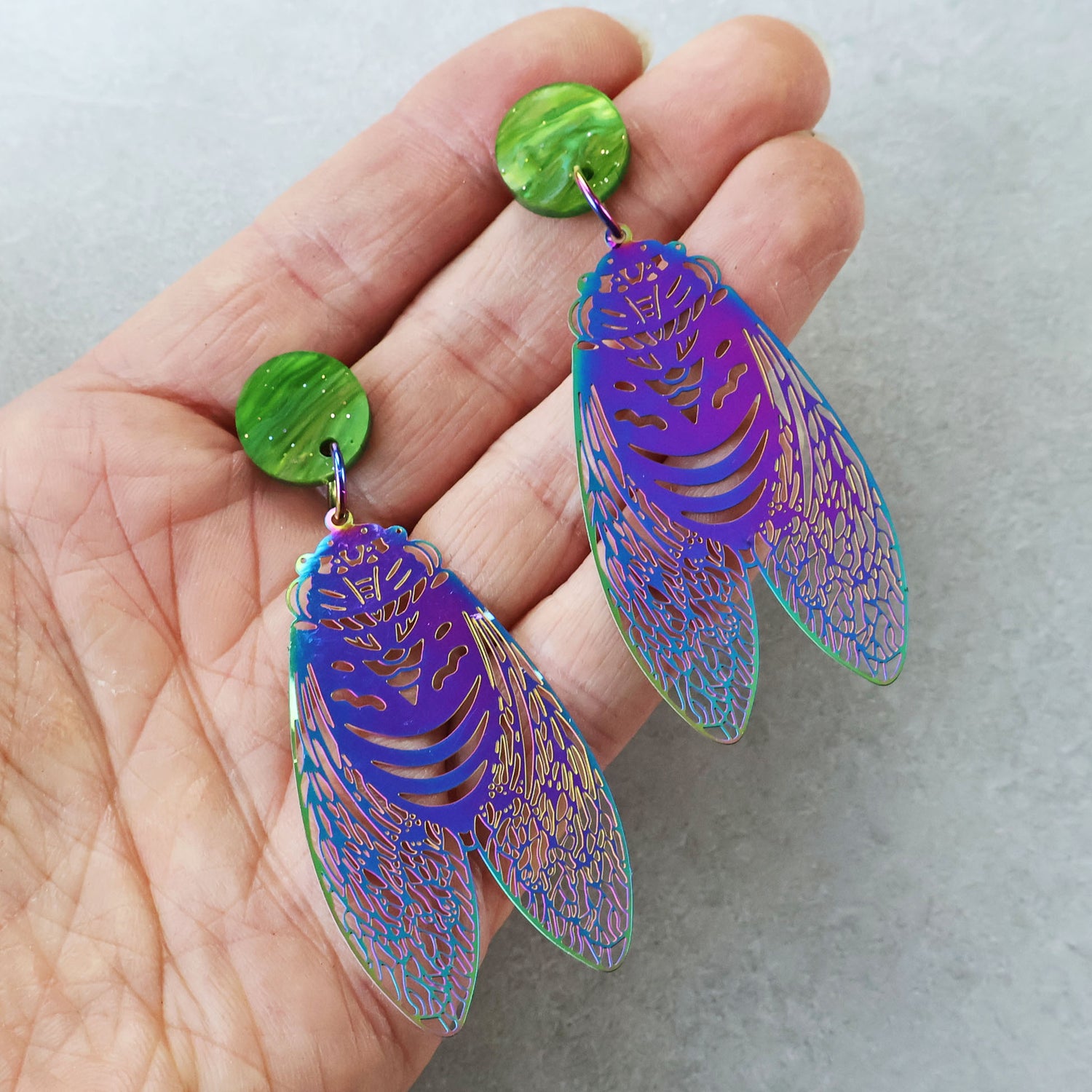 A pair of dangle earrings with stainless steel circada dangles hanging from green sparkle swirl acrylic toppers, being held in a hand. The circadas have purple/blue tones.