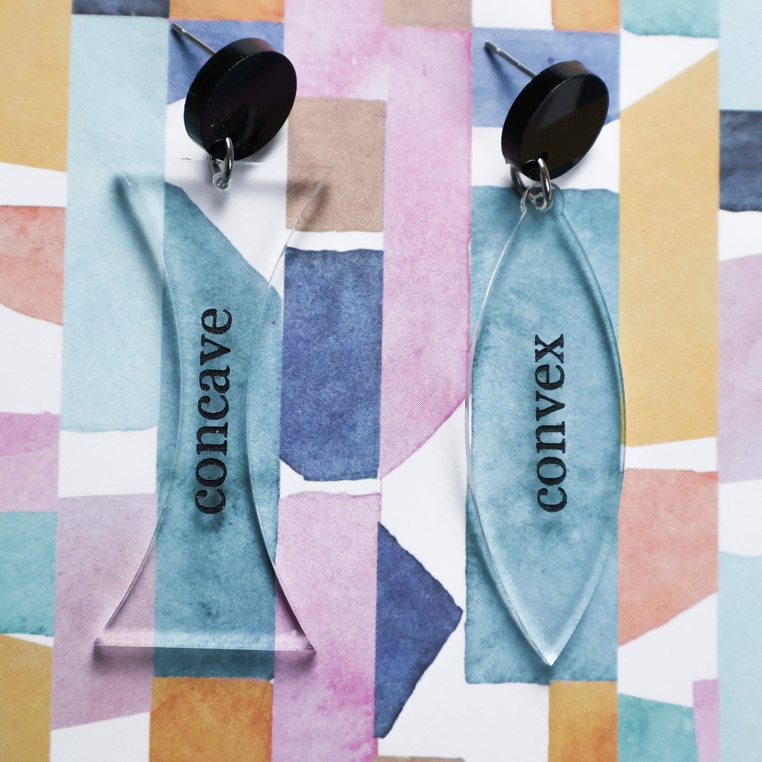 Laser cut lens shaped acrylic earrings labeled 'concave' and 'convex' against a colorful geometric pattern. Dangles are clear acrylic, text is black. Dangles hang from black round toppers. Physics themed jewellery.