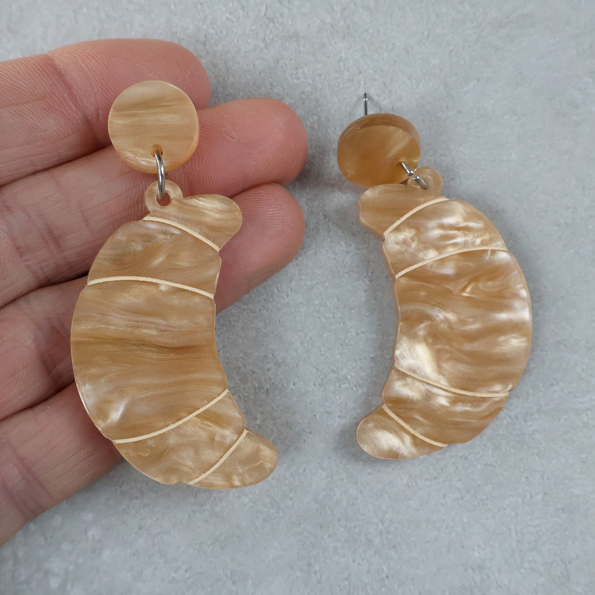 Pair of beige marbled croissant earrings held by a hand on a light grey background