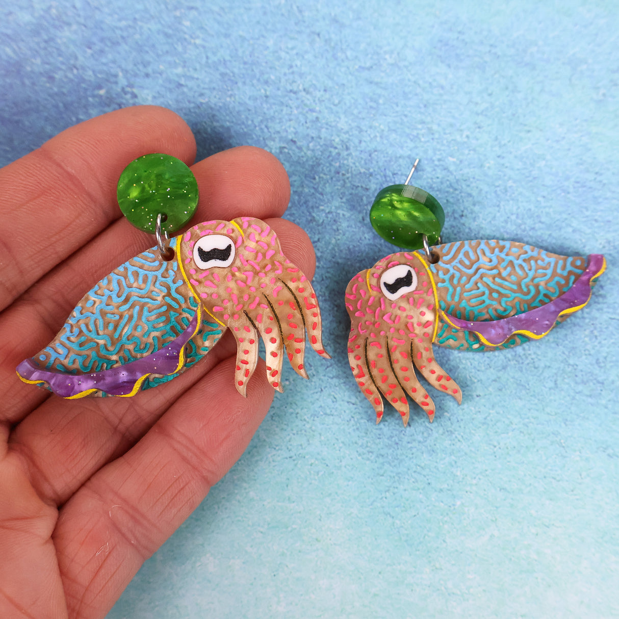 Colorful acrylic cuttlefish-shaped earrings held by a hand on a light blue background