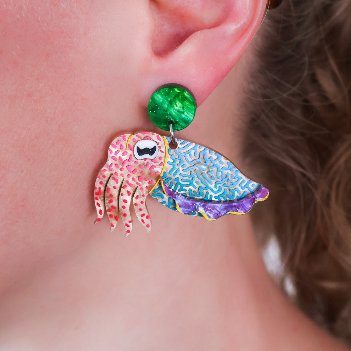 Colorful earrings with a cute cuttlefish design and green round toppers worn by a person.