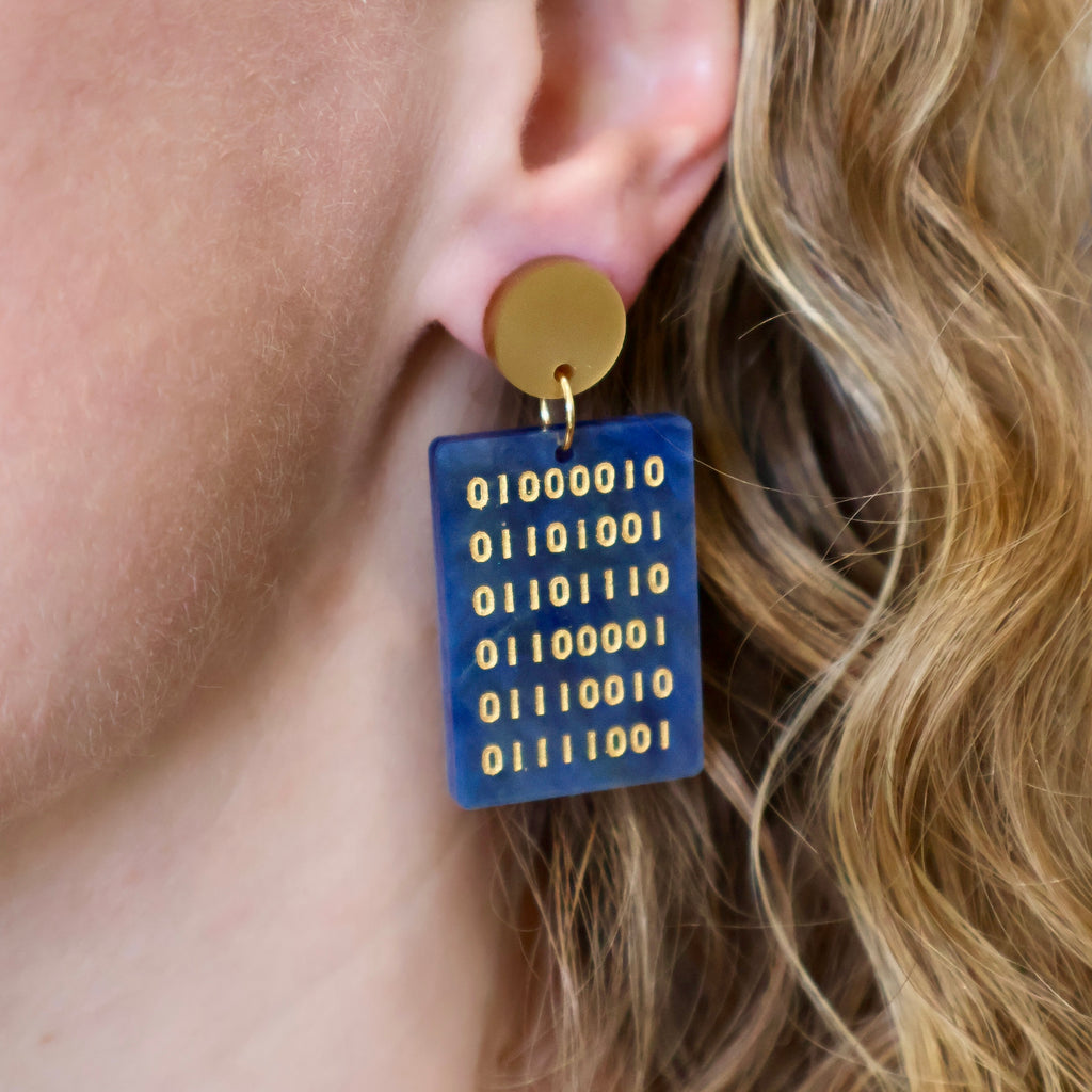 Rectangular earrings in swirly blue acrylic, with an array of gold binary code engraved within. They hang from gold toned acrylic toppers. Earrings are being modelled. 