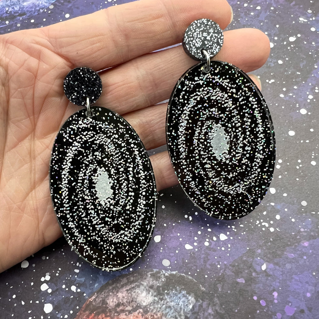 Statement sized milky way galaxy acylic earrings. The earrings are oval in shape and contain silver toned stars handpainted against a black background. The earrings hang from black glitter acrylic toppers. Earrings are being held up for a better view. 