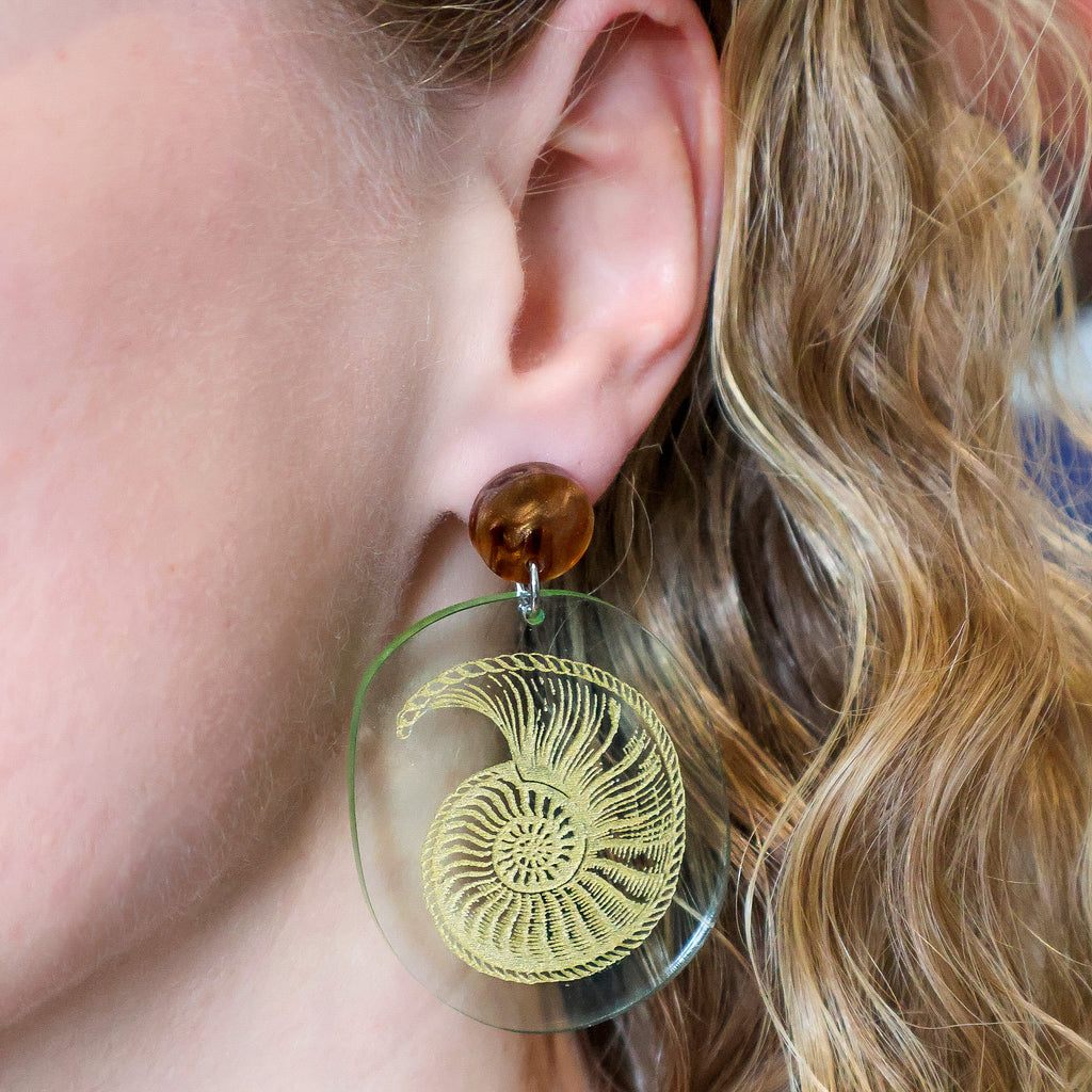 Nautilus fossil earrings, with a gold nautilus design handpainted on a green seaglass toned acrylic. They earrings hang from brown acrylic toppers. Earrings are being modelled.