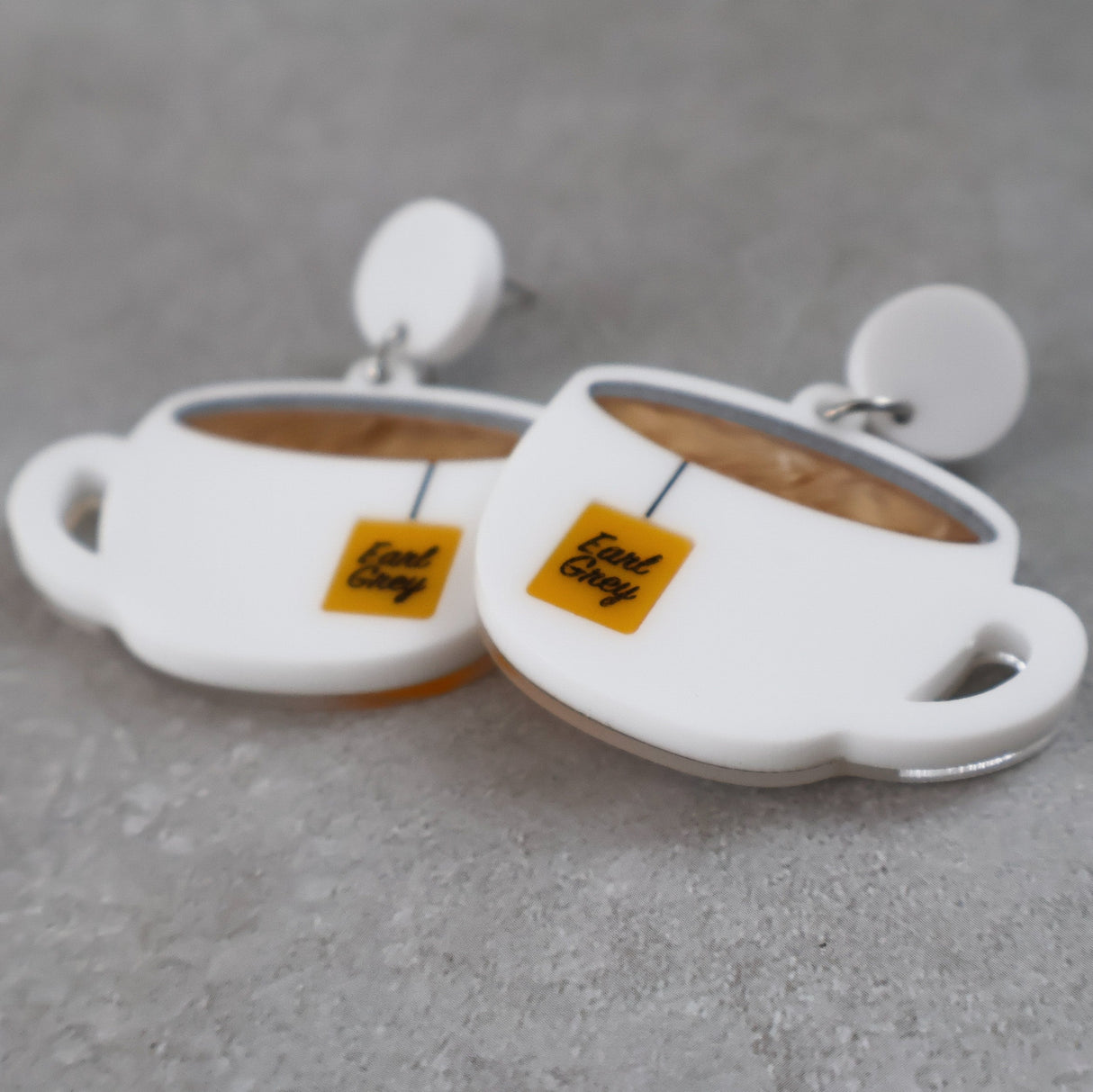 White tea cup acrylic earrings with Earl Grey tea bag tags against a gray background