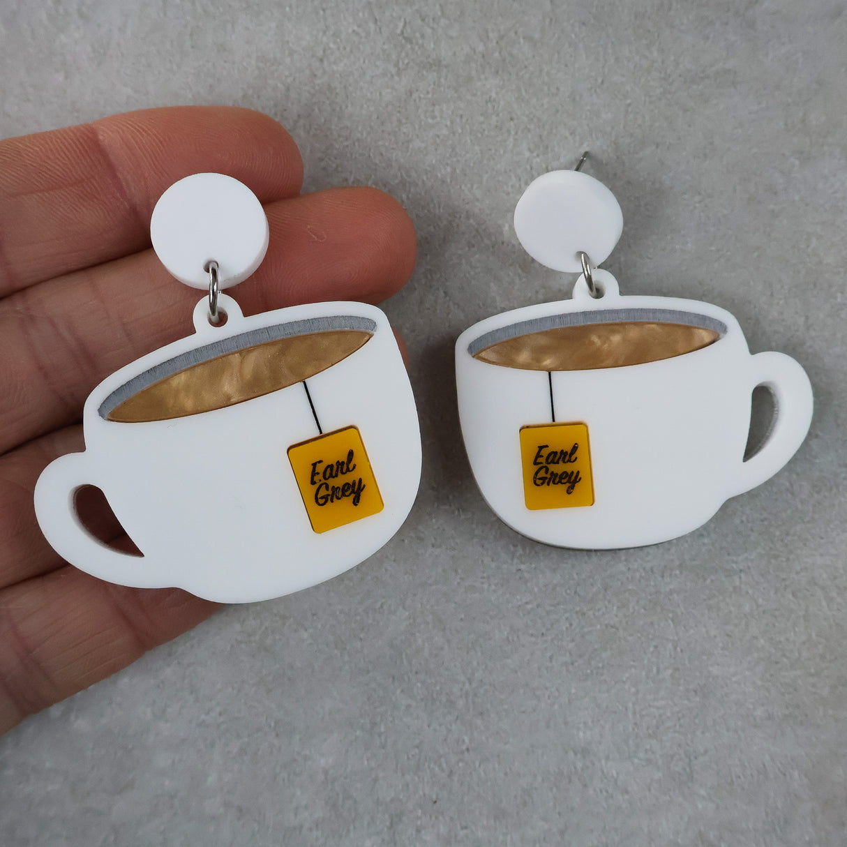 Teacup earrings with 'Earl Grey' tags held by a hand on a gray background