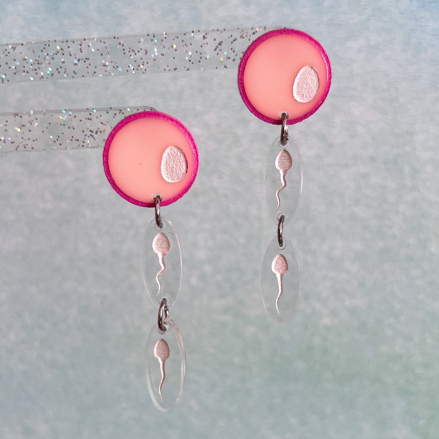 A pair of egg and sperm earrings. Two sperm dangle below a pink acrylic 'egg' topper.