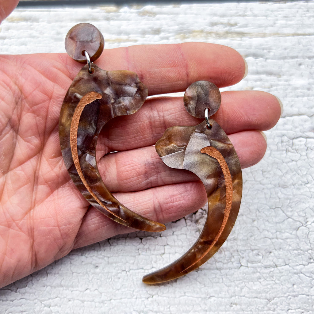 Velociraptor claw earrings, laser cut from textured brown acrylic, and hanging from brown acrylic earring toppers. Being held in hand to show scale.