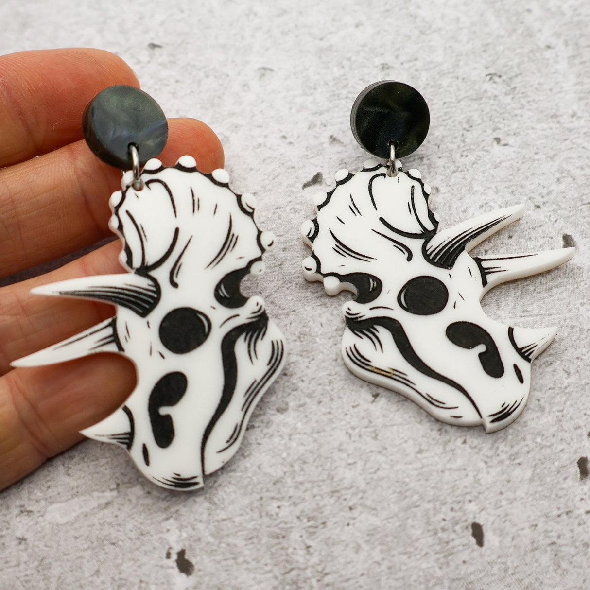 Black and white triceratops skull head acrylic earrings held by a hand on a grey surface.